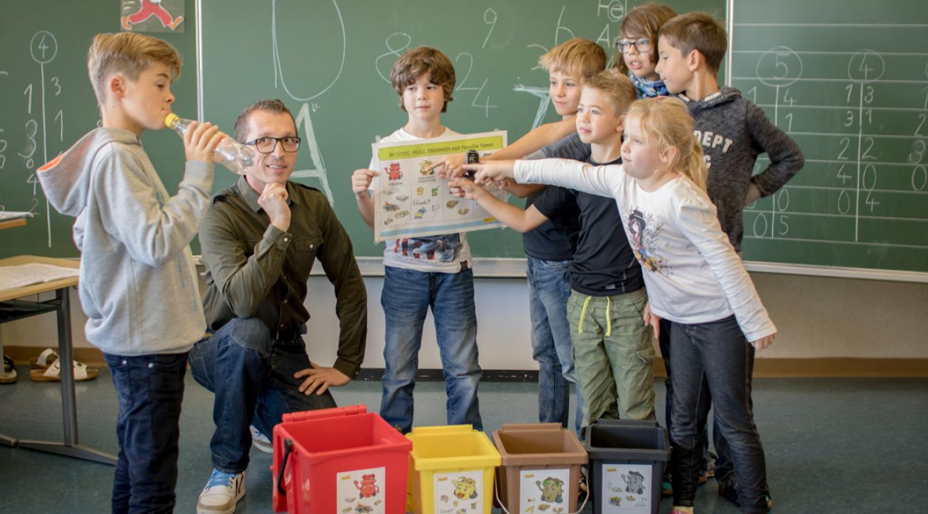 ein Abfallberater steht mit mehreren Kindern im Klassenzimmer, gemeinsam erörtern sie anhand eines Plakats die richtige Abfalltrennung