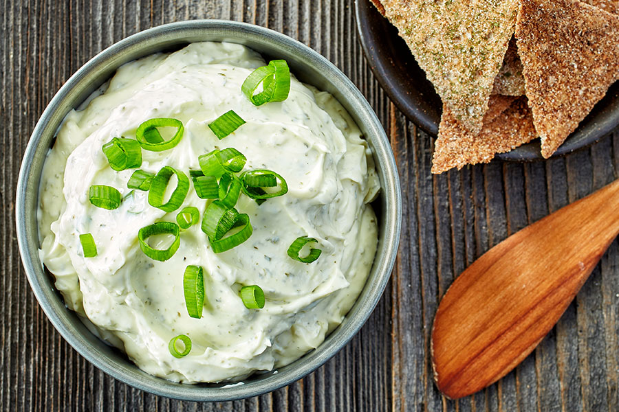 Cremiger Frühlingszwiebel-Dip in Metallschüssel, garniert mit Frühlingszwiebelringerln. Daneben ein Holzkochlöffel und eine Schüssel mit Crackern.