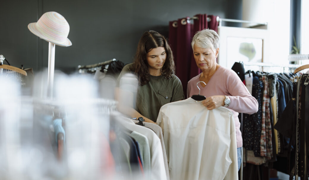 Eine ältere Dame mit grauem Haar steht in einem Secondhandladen und hält eine weiße Bluse in der Hand. Neben ihr steht ein junges Mädchen mit lockigem Haar und einem olivgrünen T-Shirt, das ebenfalls die Bluse interessiert betrachtet.