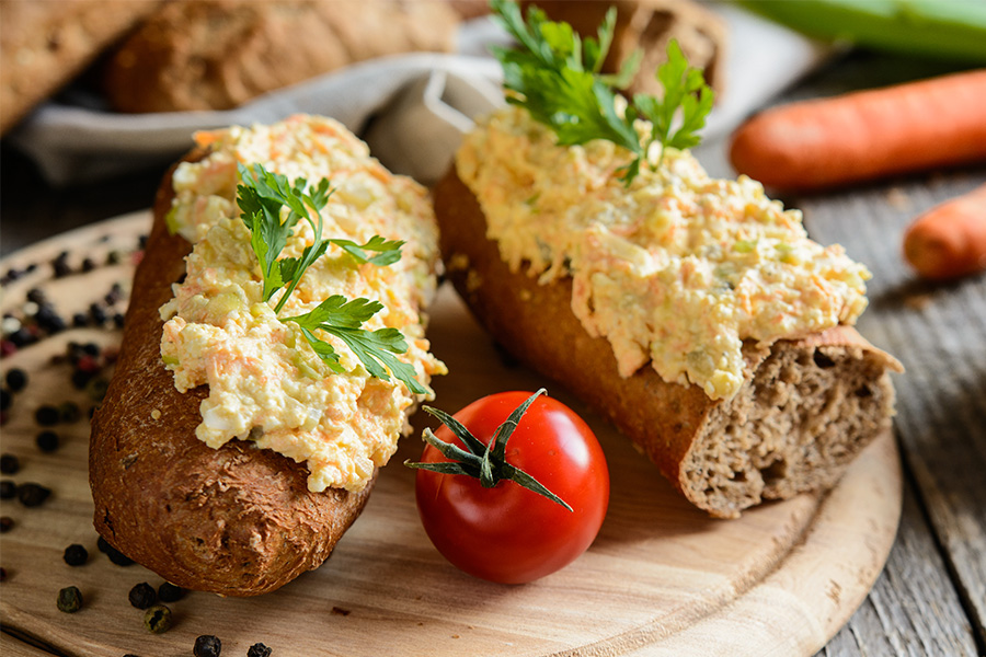 Zwei Kornspitz bestrichen mit einem Aufstrich mit geriebenen Karotten arrangiert auf einem runden Jausenbrett daneben Pfefferkörner und eine Tomate. Im Hintergrund verschwommen Karotten und Gebäck.